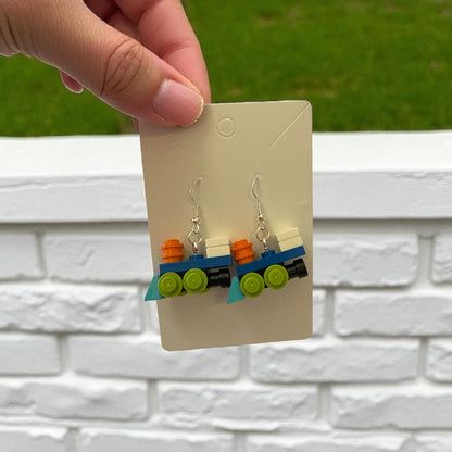 Train-shaped earrings on a display card held by a hand against a white brick wall.